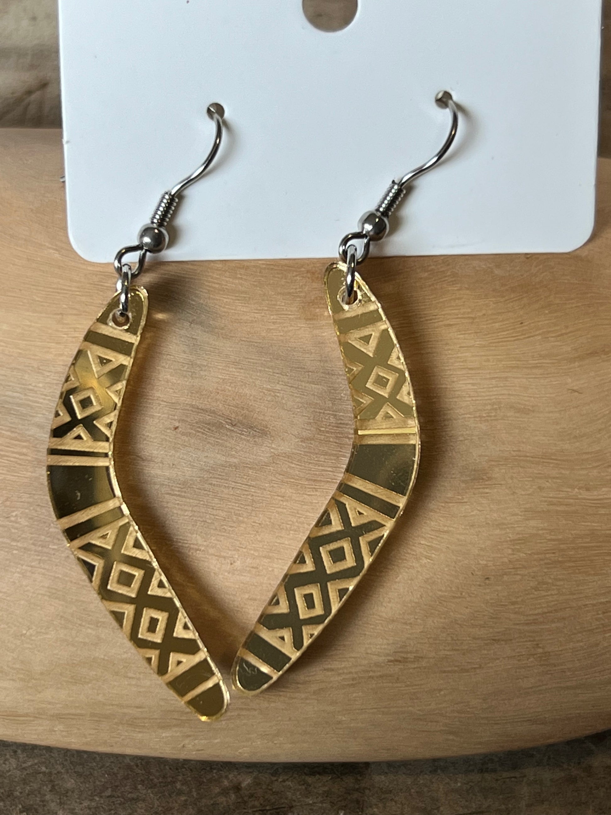 A pair of gold-colored boomerang earrings with stainless steel hooks, displayed on a cardboard hanger.