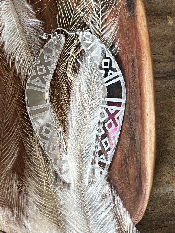 A pair of silver boomerang-shaped earrings with geometric patterns displayed on a wooden surface with a feather in the background.